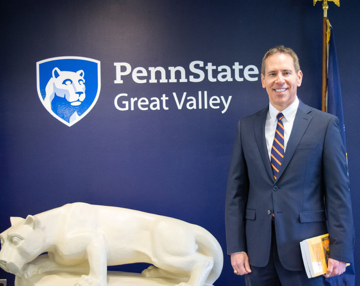 John J. Sosik standing in front of a nittany lion statue and a blue wall with the Penn State Great Valley logo