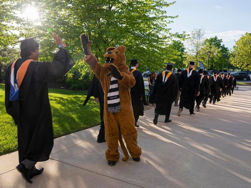 Graduates walking along the sidewalk with one high fiving the Nittany Lion