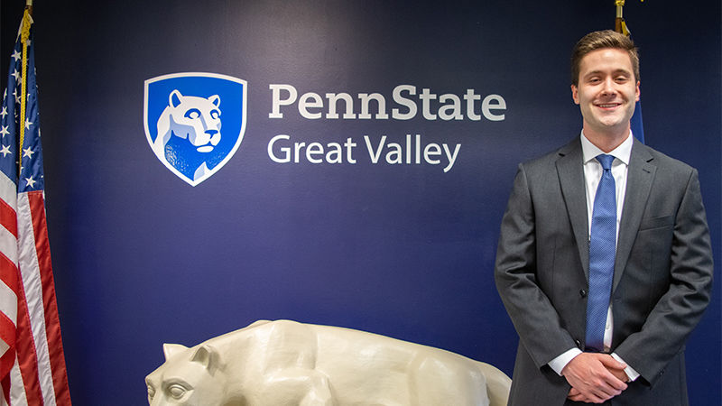 John Boland standing next to a Nittany Lion statue under the Penn State Great Valley logo