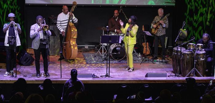 Seven musicians play a variety of instruments, from drums to trumpets to a bass, on a stage.