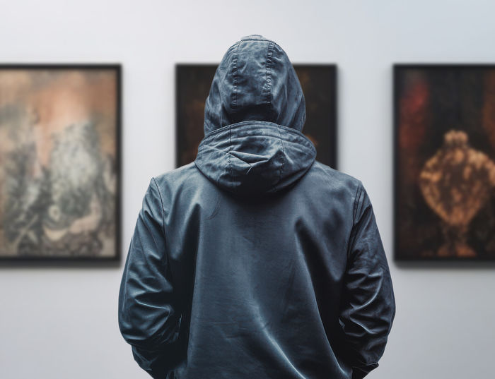 A person in a hoodie looks at three works of art on the walls.