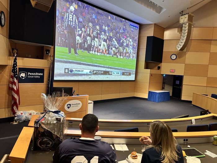 Two guests at the tailgate watch a Penn State football game in the auditorium on a large screen.