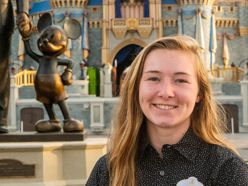 Hannah Schneidewind poses for a photo behind a Disney castle and a statue of Mickey Mouse to the left of her