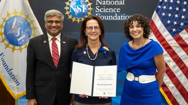Three people standing in front of a wall with the NSF logo. Kathryn Jablokow is in the middle, holding an award.