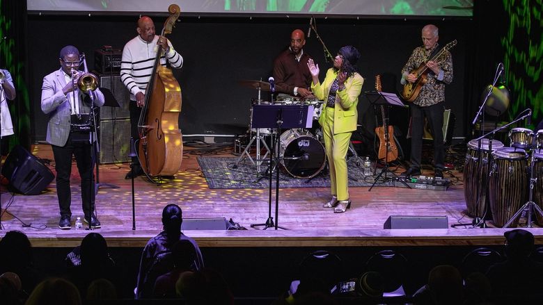 Seven musicians play a variety of instruments, from drums to trumpets to a bass, on a stage.