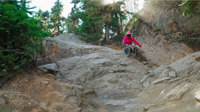An adaptive mountain biker maneuvers down a steep rocky slope below trees
