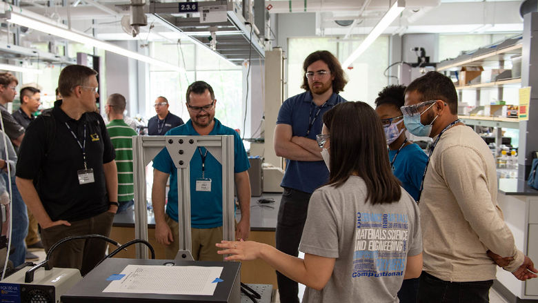 An undergraduate student gives a demo to students of the additive manufacturing and design master's program in an engineering lab with equipment on a table.