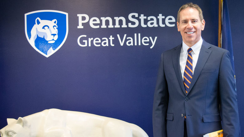 John J. Sosik standing in front of a nittany lion statue and a blue wall with the Penn State Great Valley logo