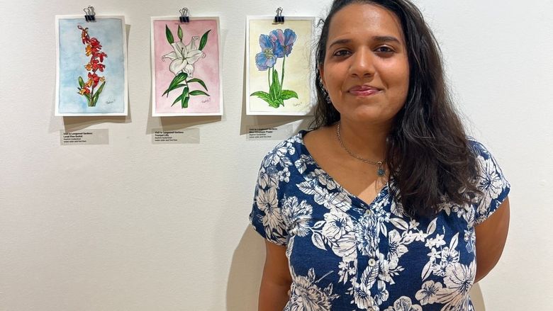 Artist poses in front of three watercolor paintings in an art exhibit