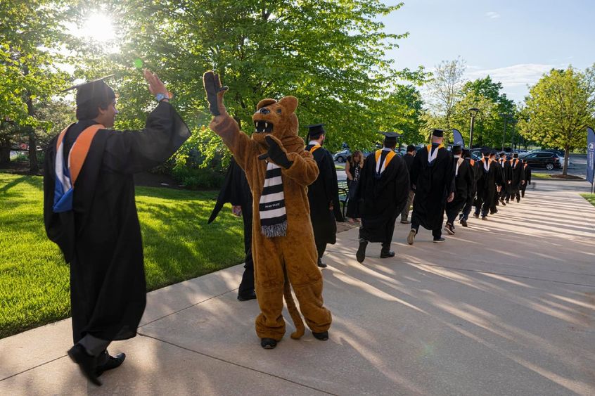 Graduates walking along the sidewalk with one high fiving the Nittany Lion