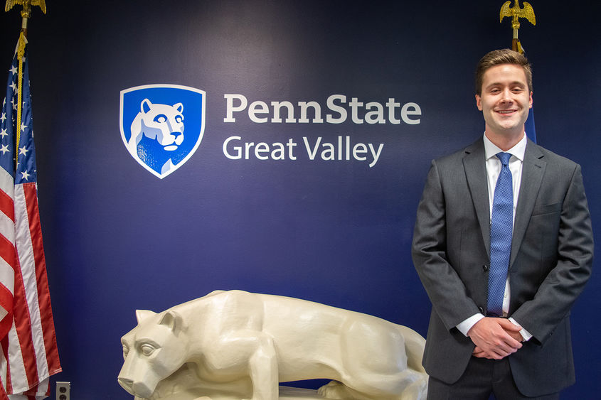 John Boland standing next to a Nittany Lion statue under the Penn State Great Valley logo